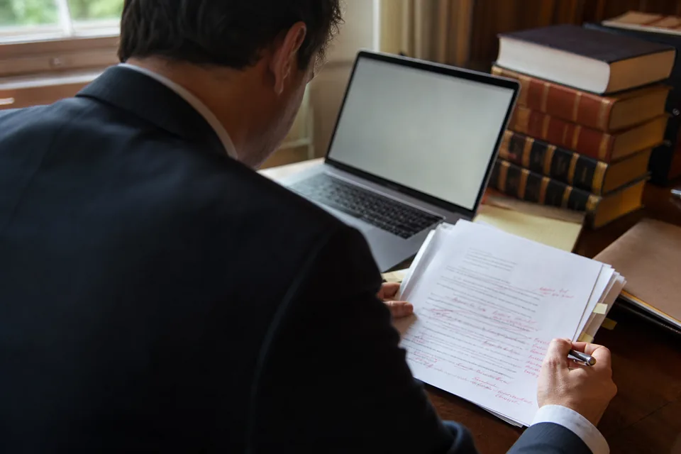Attorney seen from behind editing a printed brief with red pen marks beside a laptop and case reporters