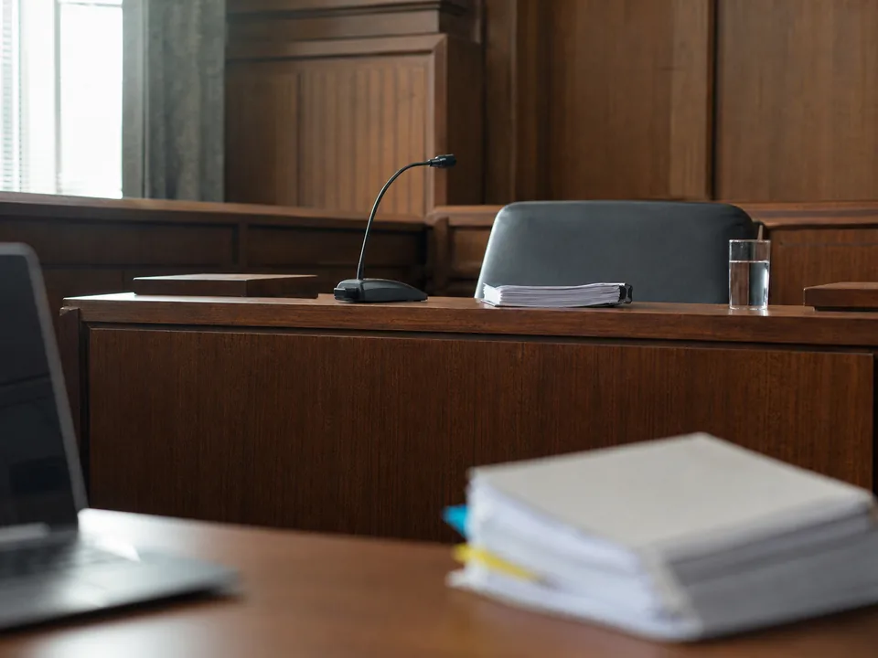 Empty courtroom witness stand seen from counsel table with an open laptop and tabbed briefs